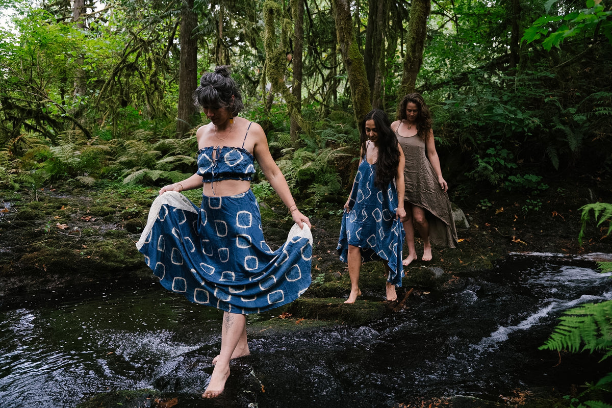 Three women in plant-dyed silk crossing a forest creek barefoot